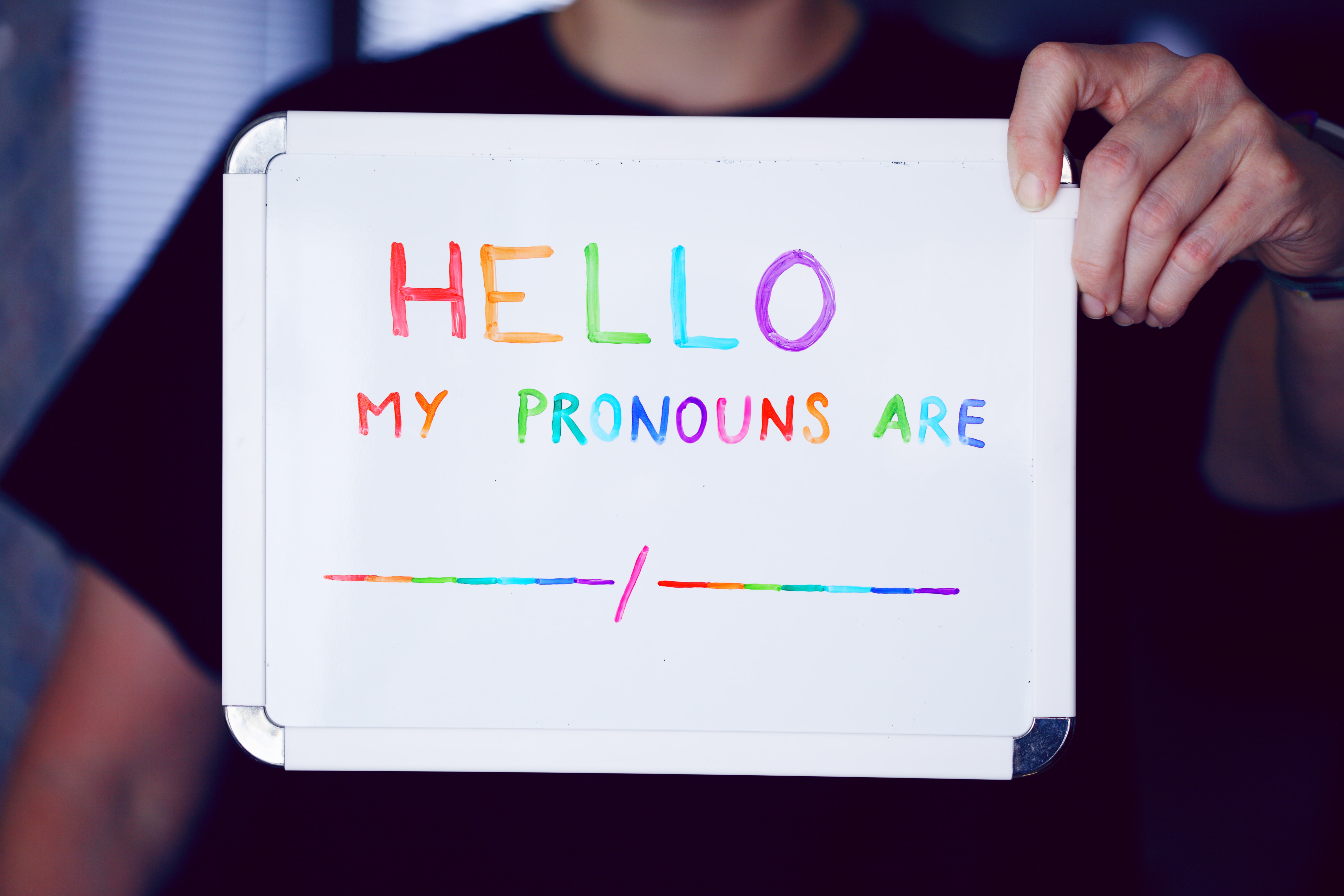 Person holding a small whiteboard with the words 'Hello my pronouns are' written in rainbow colours